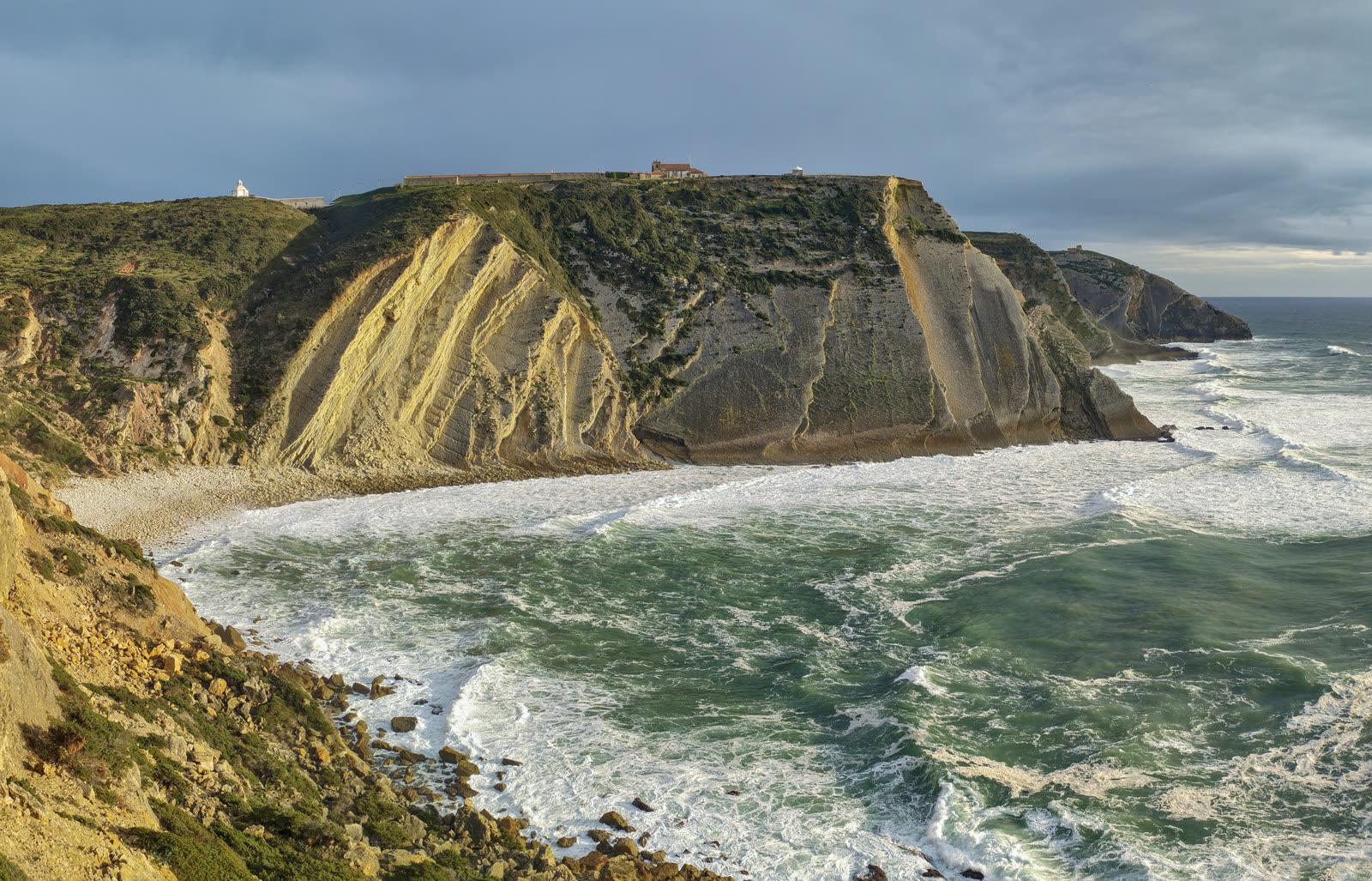 Steeply tilted pale limestone beds forming the Pedra da Mua sea cliff, with the sanctuary buildings on the plateau above and surf breaking below.