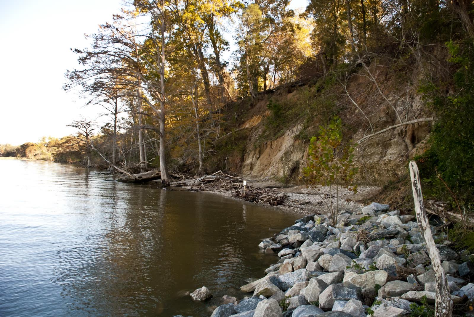 Calm James River beside a narrow beach, with granite riprap in front of a wooded bank cut back to pale sandy clay