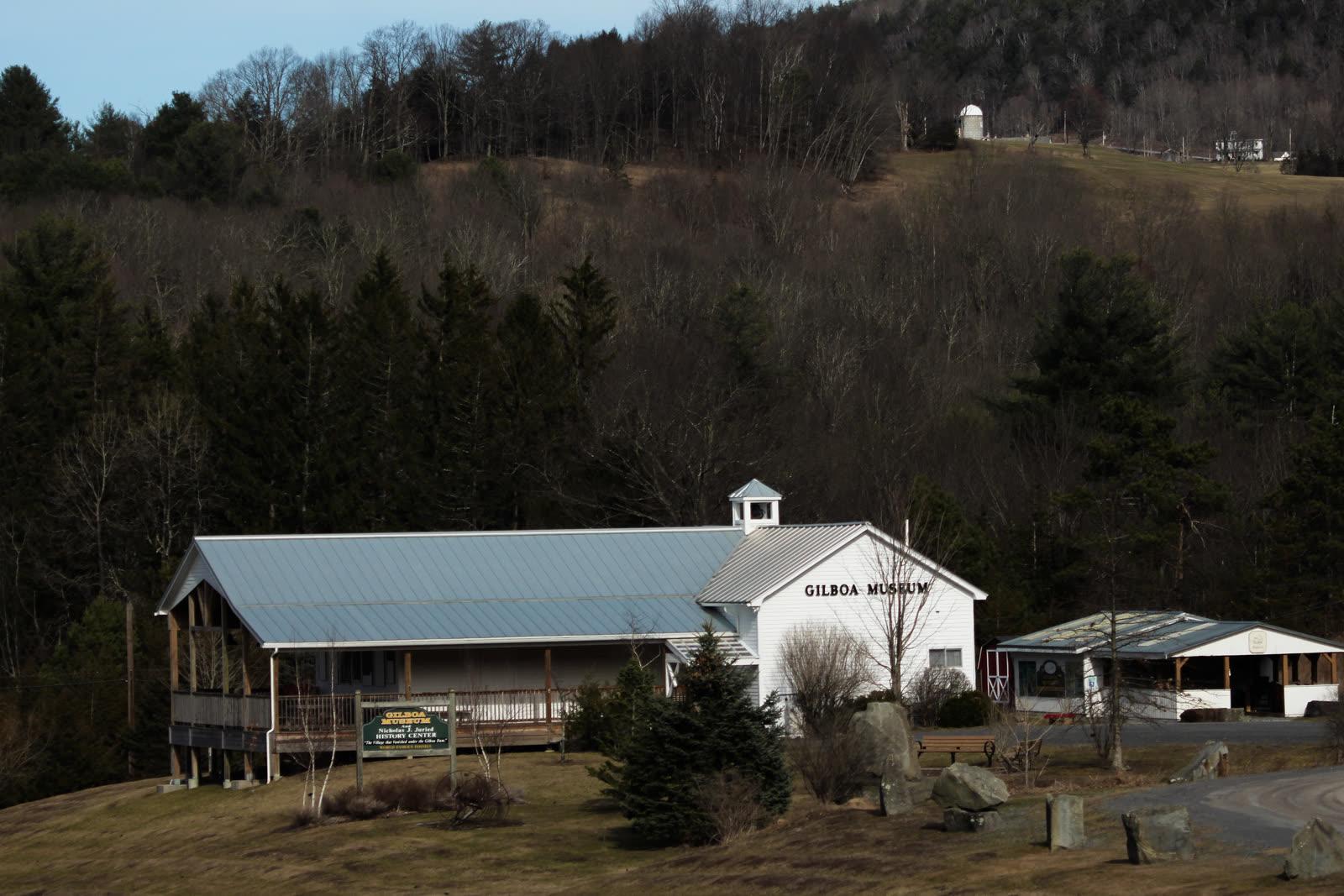 White Gilboa Museum building with a metal roof and a roadside sign, set against a wooded Catskill hillside