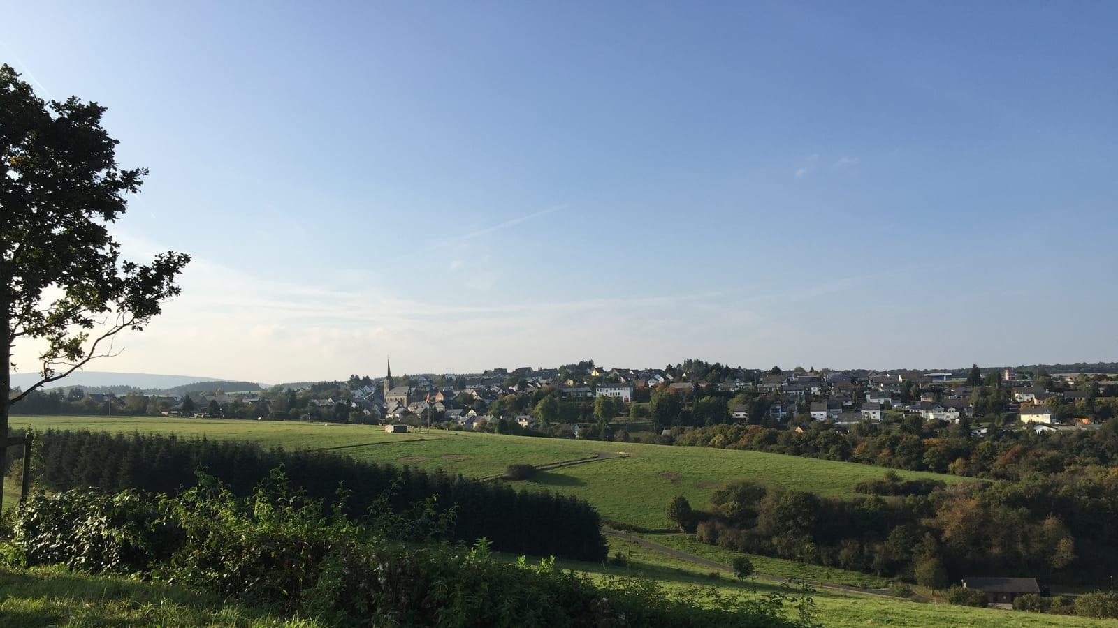 Green meadow looking across to the village of Bundenbach with a church spire and wooded hills in the distance.