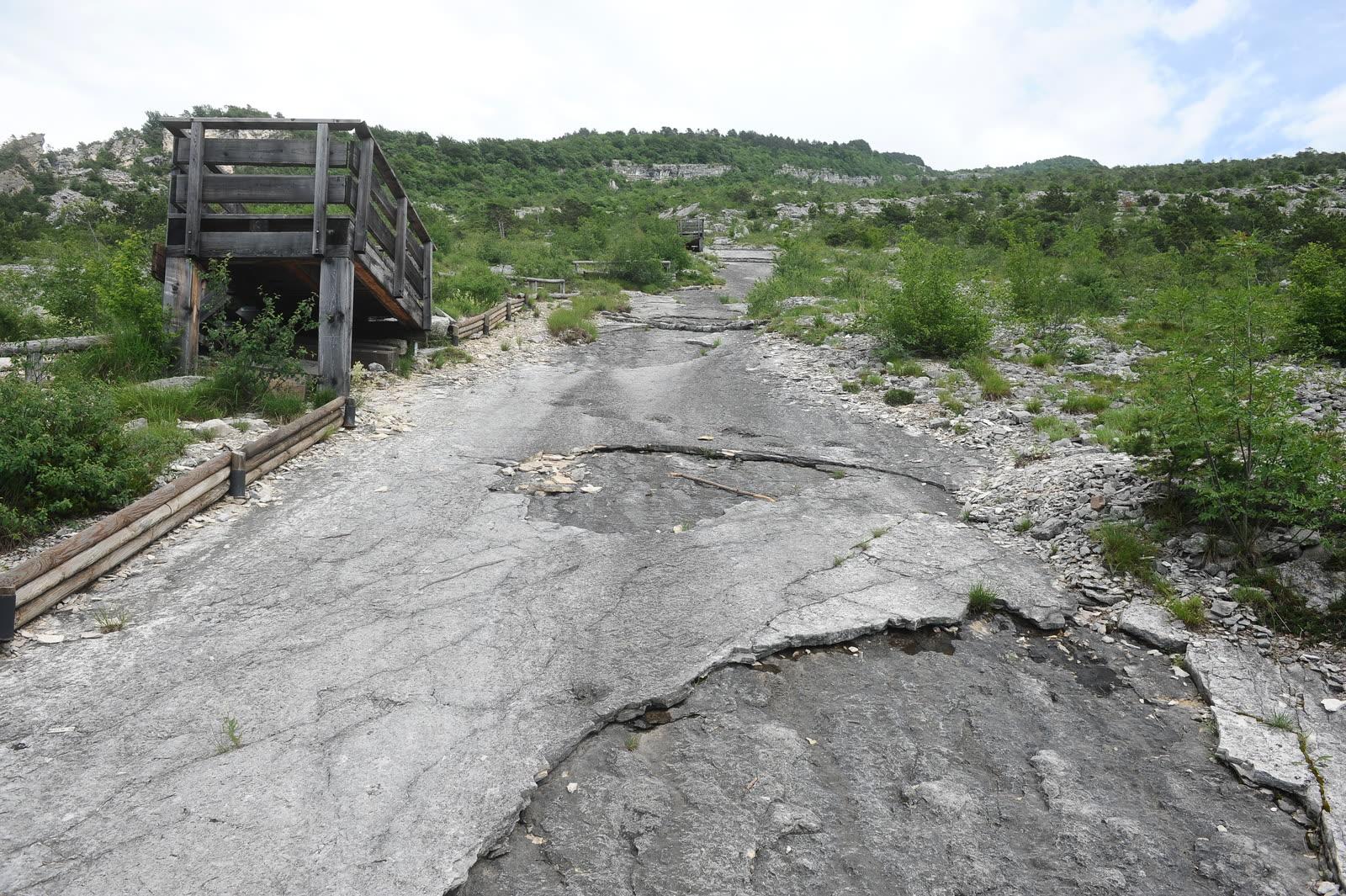 Steeply tilted pale grey limestone pavement running uphill between scrub, with a timber viewing tower and wooden rail at the left edge under a hazy sky.