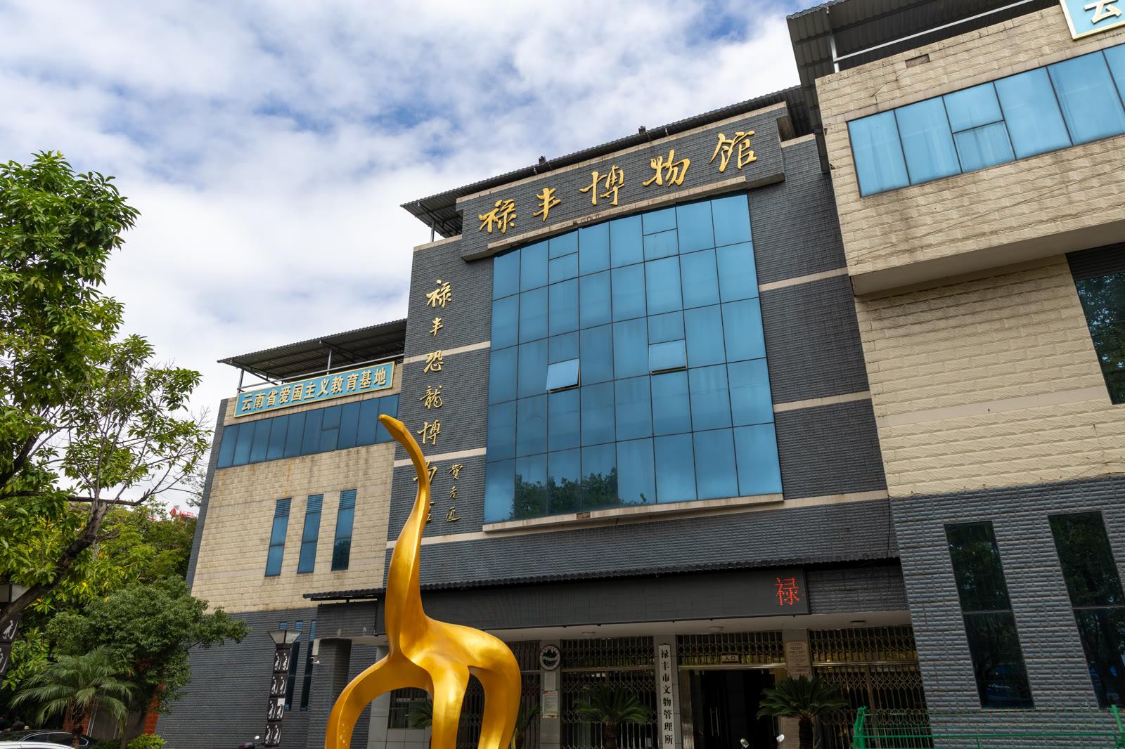 The glass and concrete frontage of the Lufeng Dinosaur Museum with a tall golden sauropod sculpture outside