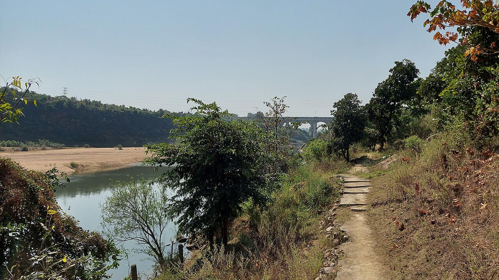 Footpath along the wooded bank of the Hasdeo River at the Gondwana fossil park, with a rail viaduct in the distance