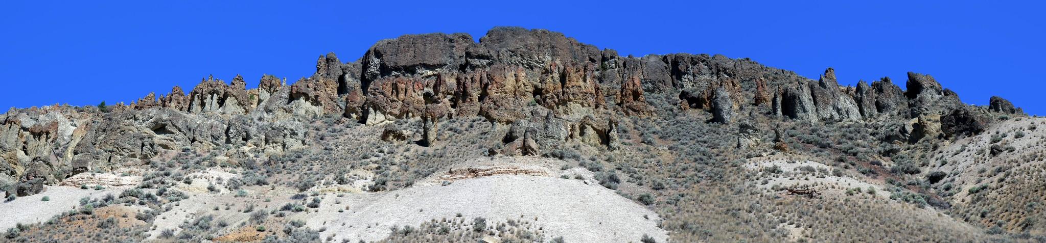 Panorama of a ridge of weathered basalt hoodoos above a pale grey shale slope under a clear blue sky near Cache Creek, British Columbia