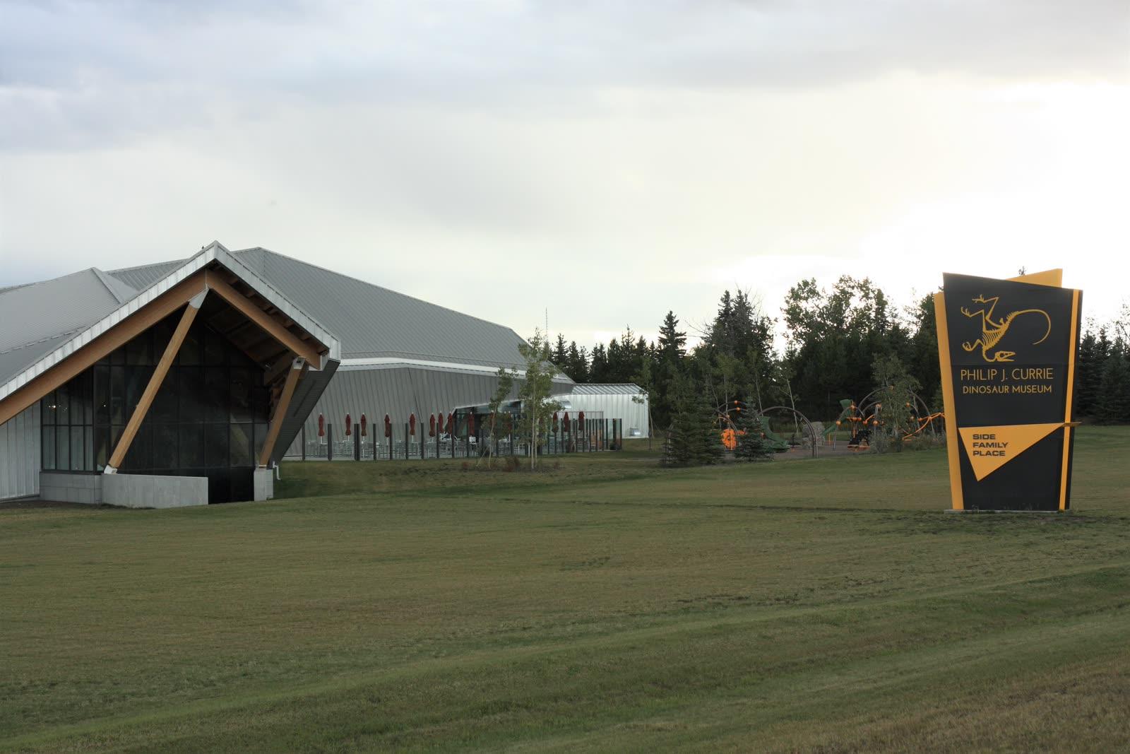 Timber-framed glass and metal exhibition hall of the Philip J. Currie Dinosaur Museum beside its yellow roadside sign in Wembley, Alberta
