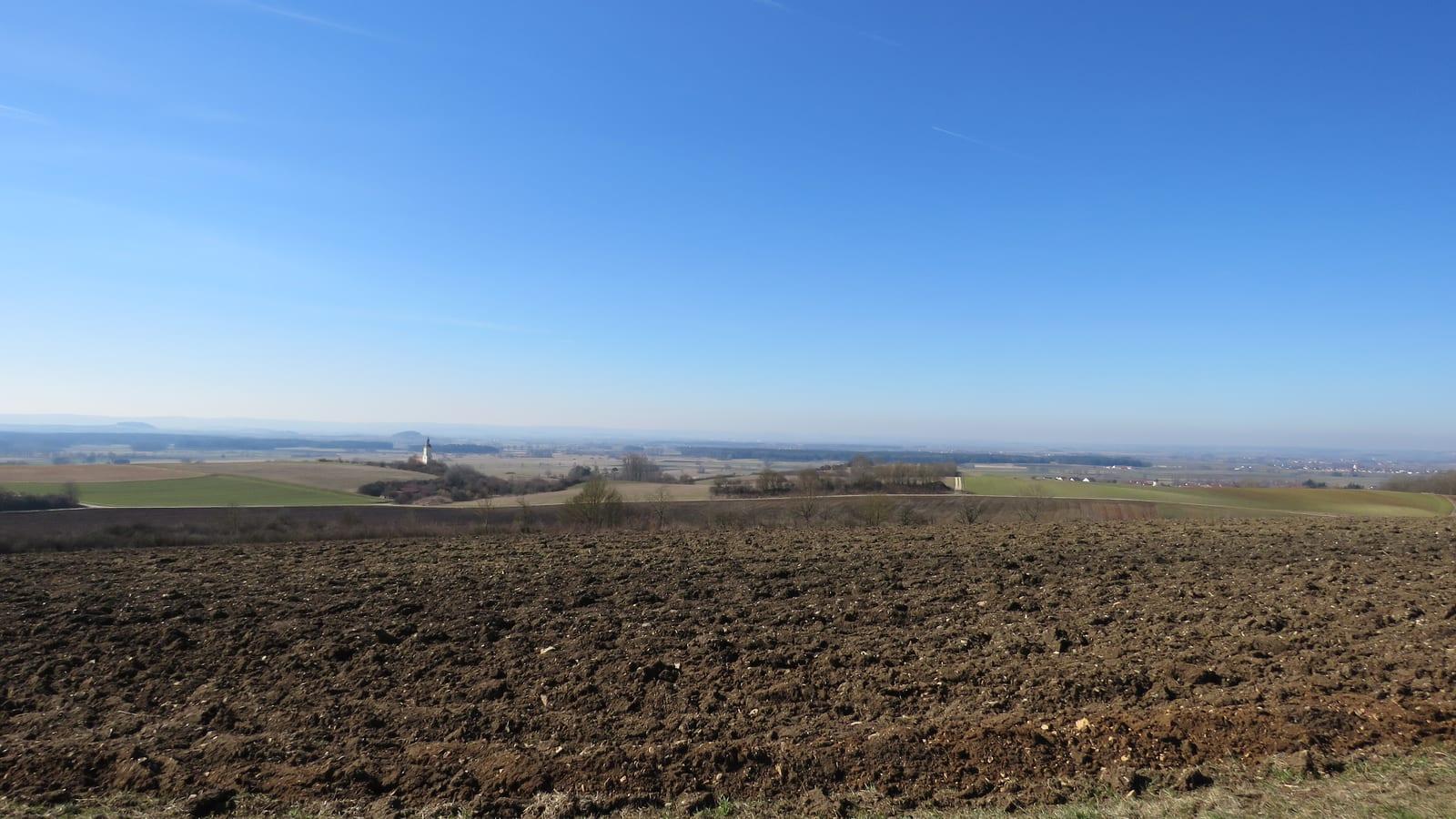 Ploughed brown field with a distant church tower and gentle rolling farmland on the Ries Crater plateau, Bavaria.