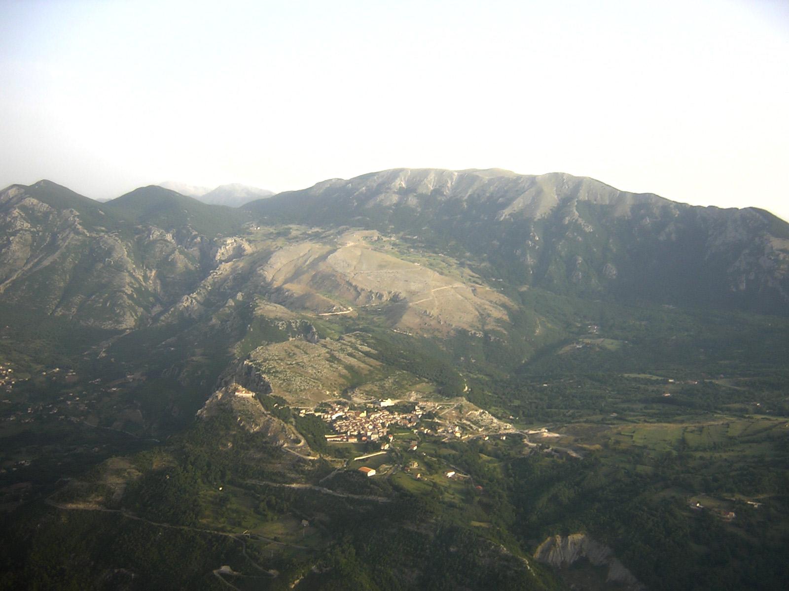 Aerial view of an Apennine village set in a green valley below a long ridge with a pale quarry scar, Campania.