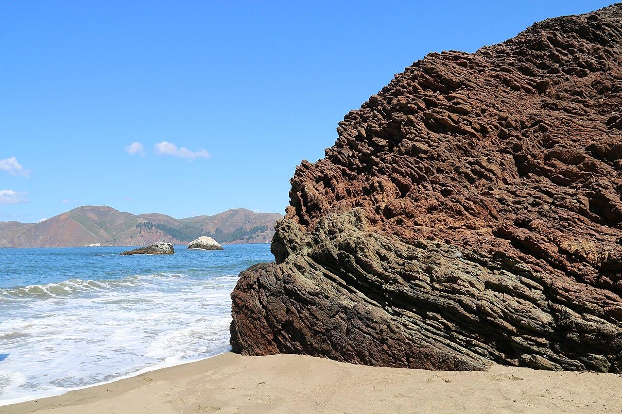 Folded red ribbon chert of the Marin Headlands rising straight out of the surf on a rocky shore
