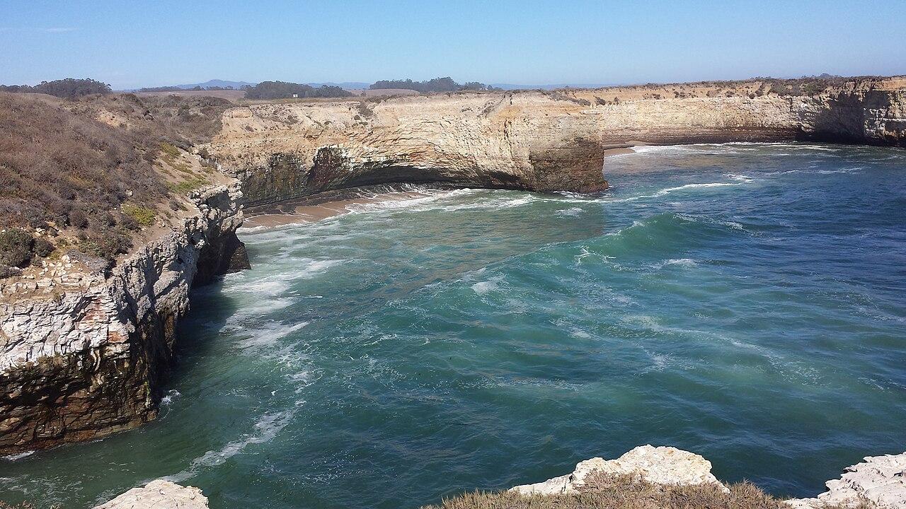 A sea arch cut through a low headland, waves washing through the opening onto a rocky Californian shore