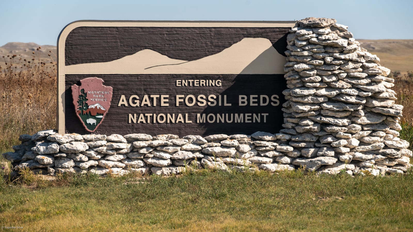 Park entrance sign reading 'Entering Agate Fossil Beds National Monument' set on a dry-stacked stone wall.