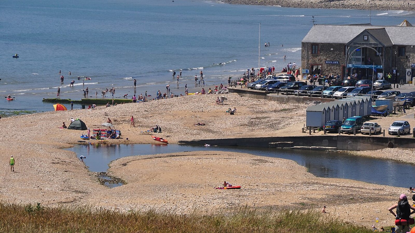 Crowded shingle beach with families and the Heritage Centre building above the shore at Charmouth, Dorset.
