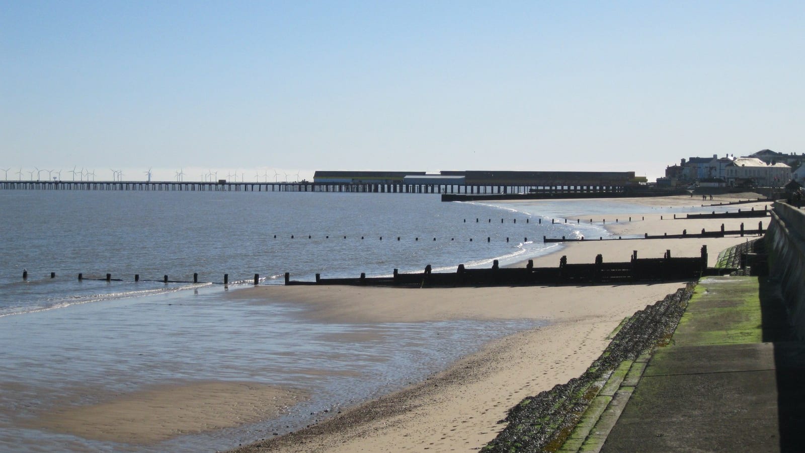 Wide sandy beach with timber groynes at low tide and a distant pier and wind farm at Walton-on-the-Naze, Essex.