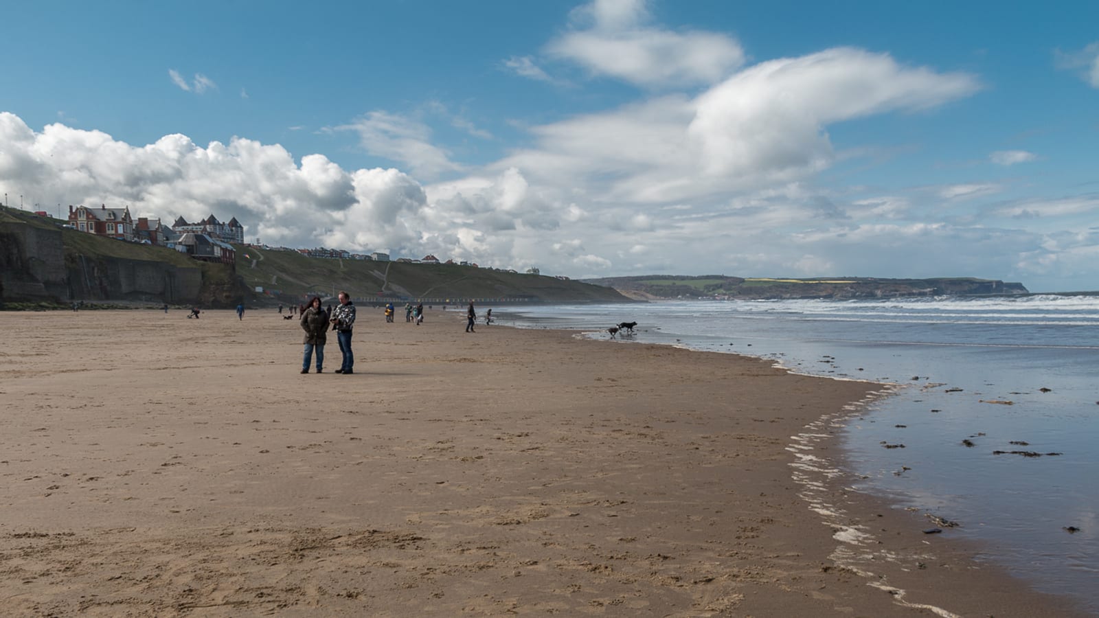Wide sandy beach with dog walkers and the West Cliff hotels above the sand at Whitby on a bright cloudy day.