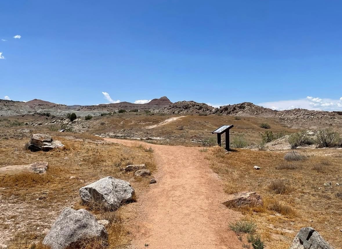 Packed sand trail through open desert at the Fruita Paleontological Area, with an interpretive panel standing beside it and banded badlands on the skyline.
